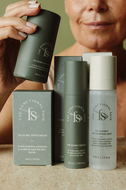 A close-up of a person holding a dark green Functional Skin cosmetic container, with several Functional Skin skincare products arranged on a countertop in front of them, including a boxed moisturiser, a serum bottle, and a mist bottle.