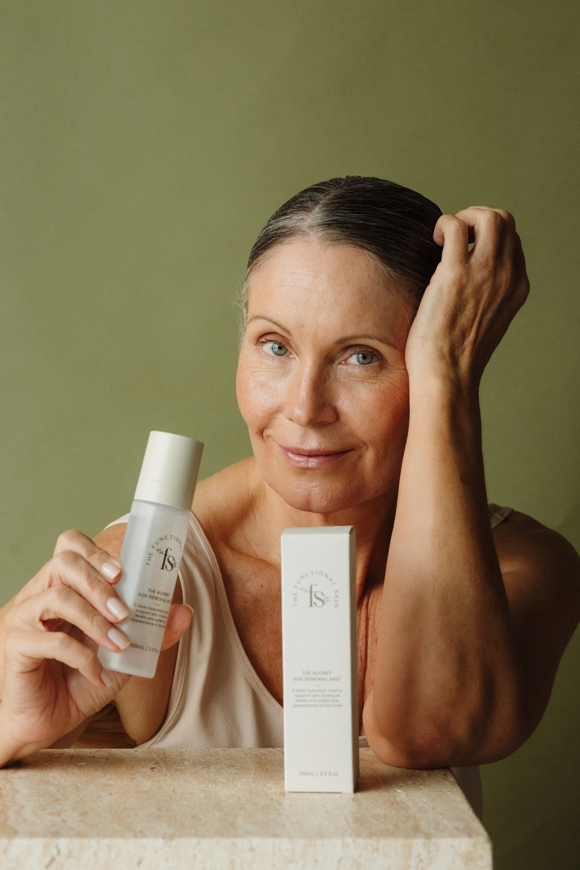 Woman holding The Audrey Age Renewal Mist against a green background